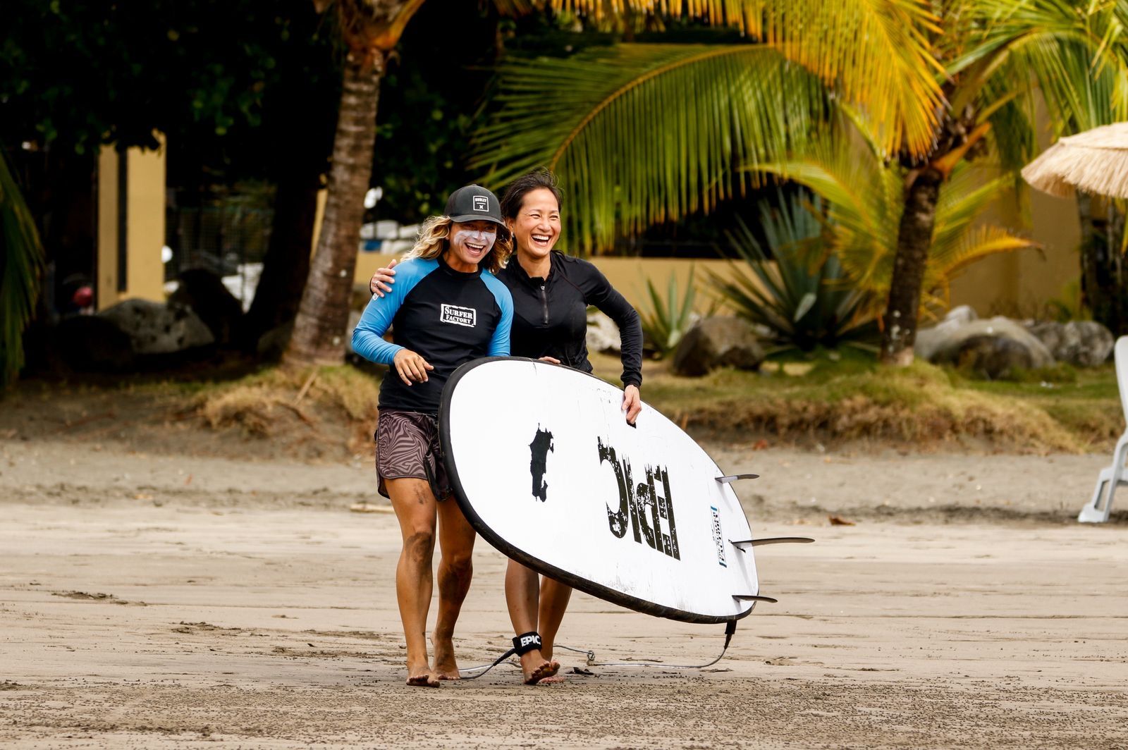 Two surfers laughing on the beach with a board